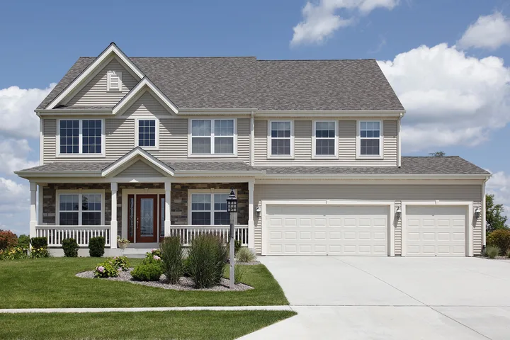 Two-story suburban home with beige siding, stone accents, a covered front porch, and a three-car garage. The property features landscaped garden beds, a manicured lawn, and a wide concrete driveway beneath a bright blue sky with scattered clouds.