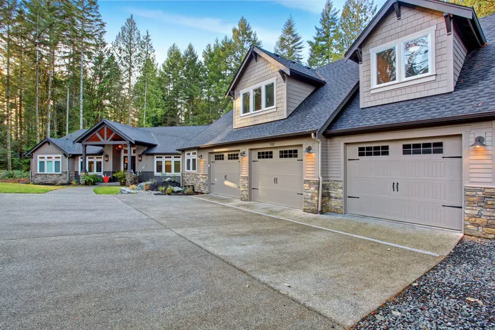 Large craftsman-style home with three-car garage, stone accents, and gray siding, surrounded by tall evergreen trees. A wide concrete driveway leads to the covered front entrance, showcasing the home's spacious exterior and wooded setting.